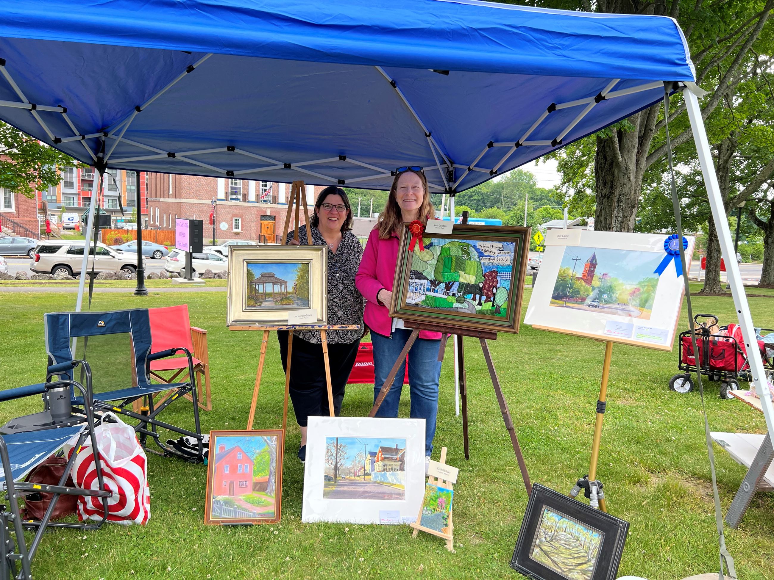 two woman standing with a group of paintings and quilts